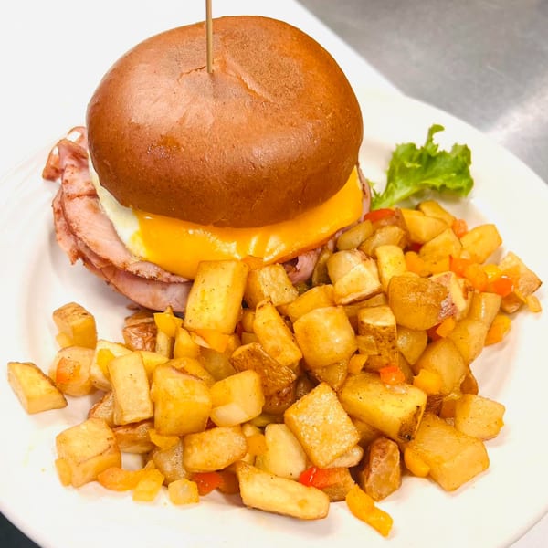 A breakfast sandwich with ham, egg, and cheese on a bun, served with a side of home fries.
