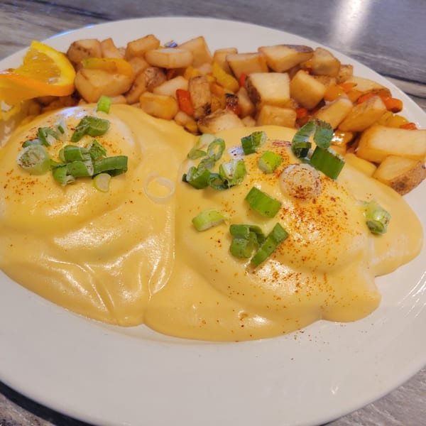 Eggs Benedict with hollandaise sauce, topped with green onions and paprika, served with home fries and an orange slice.