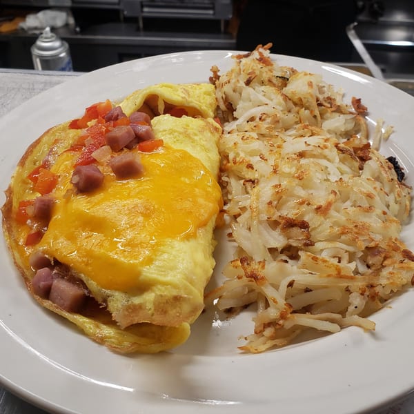 A fluffy omelet topped with melted cheese, diced ham, and red peppers, served with crispy hash browns.