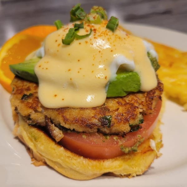 A crab cake Benedict with avocado, hollandaise sauce, and green onions on a waffle with a side of orange.