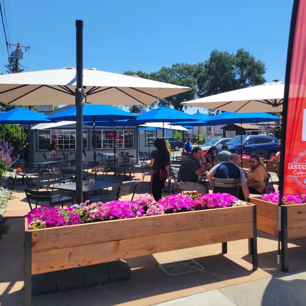 Santanas Cottage Cafe's outdoor patio with blue and white umbrellas, diners, and pink flowers in planters.