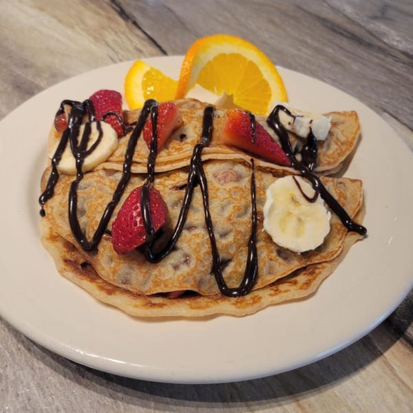 Waffle-textured pancakes topped with chocolate drizzle, sliced strawberries, bananas, and orange wedges.